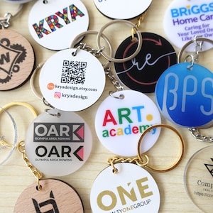 May include: A collection of circular keychains featuring various designs, including logos, QR codes, and text such as KRYA, ART academy, OAR Olympia Area Rowing, and ONE. The keychains are made of white, frosted, and wood materials with metal rings.