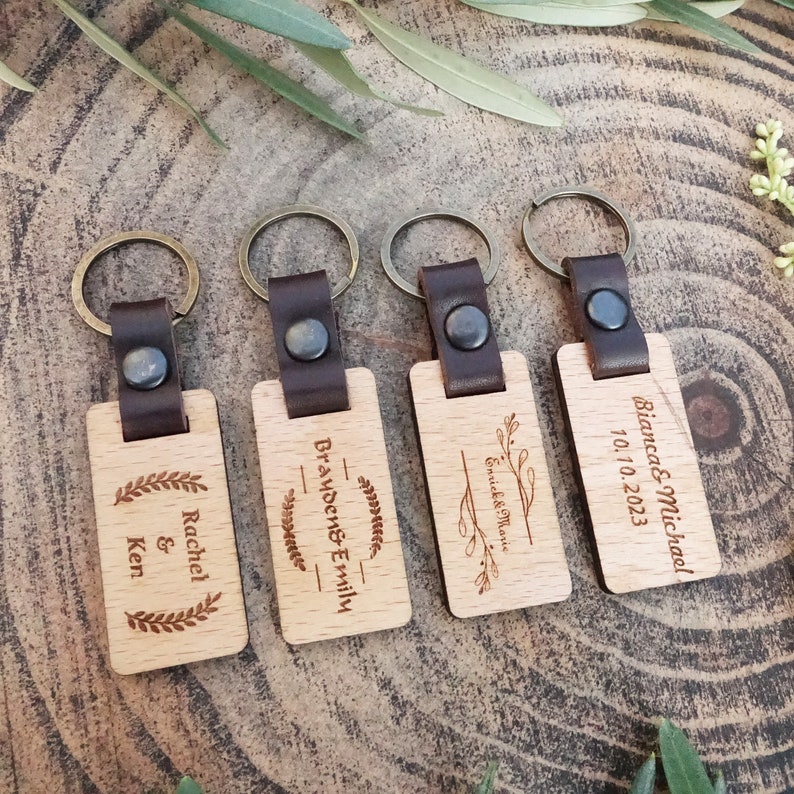 Op de afbeelding: Vier houten sleutelhangers met leren bandjes en metalen ringen. Elke sleutelhanger heeft een ander gegraveerd ontwerp met namen en data. De ontwerpen omvatten een lauwerkrans, een bloemmotief en een eenvoudig lijndesigne.