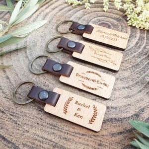 Op de afbeelding: Vier houten sleutelhangers met leren bandjes en metalen ringen. Elke sleutelhanger heeft een ander gegraveerd bericht, waaronder "Bianca & Michael 10.10.2023", "Braden & Emily", "Rachel & Ken" en "Emick & Marie".