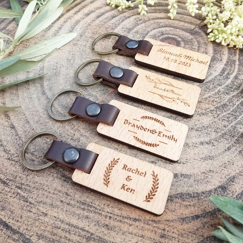 Op de afbeelding: Vier houten sleutelhangers met leren bandjes en metalen ringen. Elke sleutelhanger heeft een ander gegraveerd bericht, waaronder "Bianca & Michael 10.10.2023", "Braden & Emily", "Rachel & Ken" en "Emick & Marie".