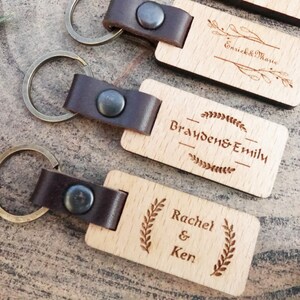Op de afbeelding: Drie houten sleutelhangers met leren bandjes en metalen ringen. Elke sleutelhanger heeft een andere naam gegraveerd: "Brayden & Emily", "Rachel & Ken" en "Enrick & Marie".