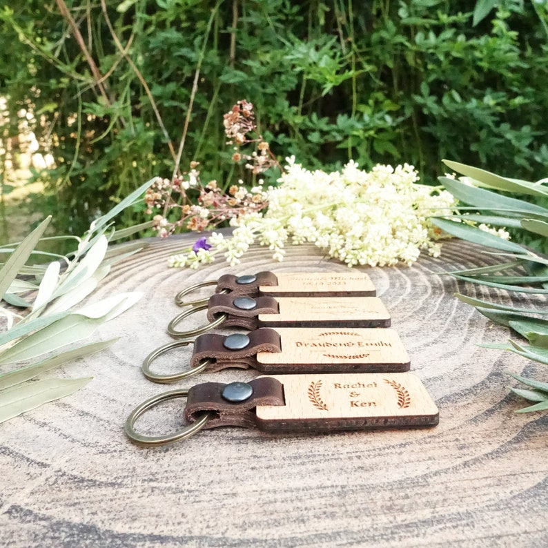 Op de afbeelding: Vier houten sleutelhangers met leren bandjes en metalen ringen. De sleutelhangers zijn gegraveerd met namen en data. De sleutelhangers zijn in een rij op een houten oppervlak gerangschikt.