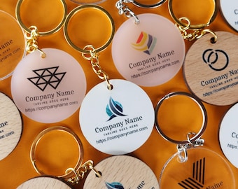 May include: A collection of circular keychains featuring various logos and the text Company Name, Tagline Goes Here, and https://companyname.com. The keychains are made of clear acrylic, frosted acrylic, and light wood, each attached to a gold metal ring.