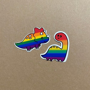 Könnte beinhalten: Zwei regenbogenfarbene Dinosaurier-Aufkleber. Einer ist ein Triceratops und der andere ein Dinosaurier mit langem Hals.