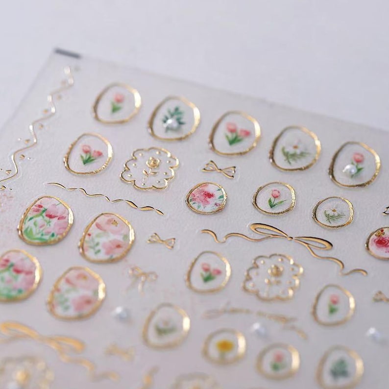 May include: A sheet of decorative stickers featuring floral designs and gold accents. The stickers include various flower illustrations, bows, and gold outlines. The stickers are in a variety of shapes and sizes, with a clear background.