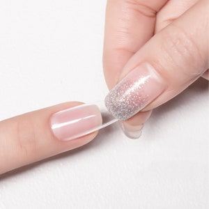 Op de afbeelding: Een hand die een heldere nagellijm aanbrengt op een vingernagel met zilveren glitternagellak.