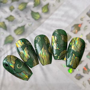 May include: Five green nail designs with leaf patterns and gold accents. The nails have a glossy finish and are arranged on a white surface. Leaf-shaped nail art stickers are visible in the background.