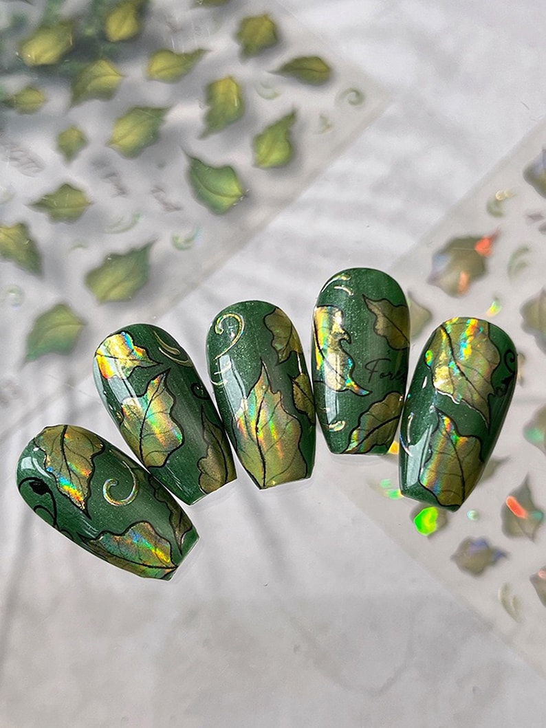 May include: Five green nail designs with leaf patterns and gold accents. The nails have a glossy finish and are arranged on a white surface. Leaf-shaped nail art stickers are visible in the background.