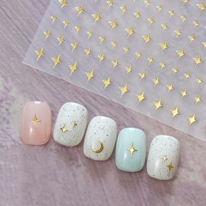 May include: A sheet of gold star nail art stickers next to five manicured nails. The nails are painted in various colours, including pink, white with speckles, and light blue. Each nail features a gold star or moon design.
