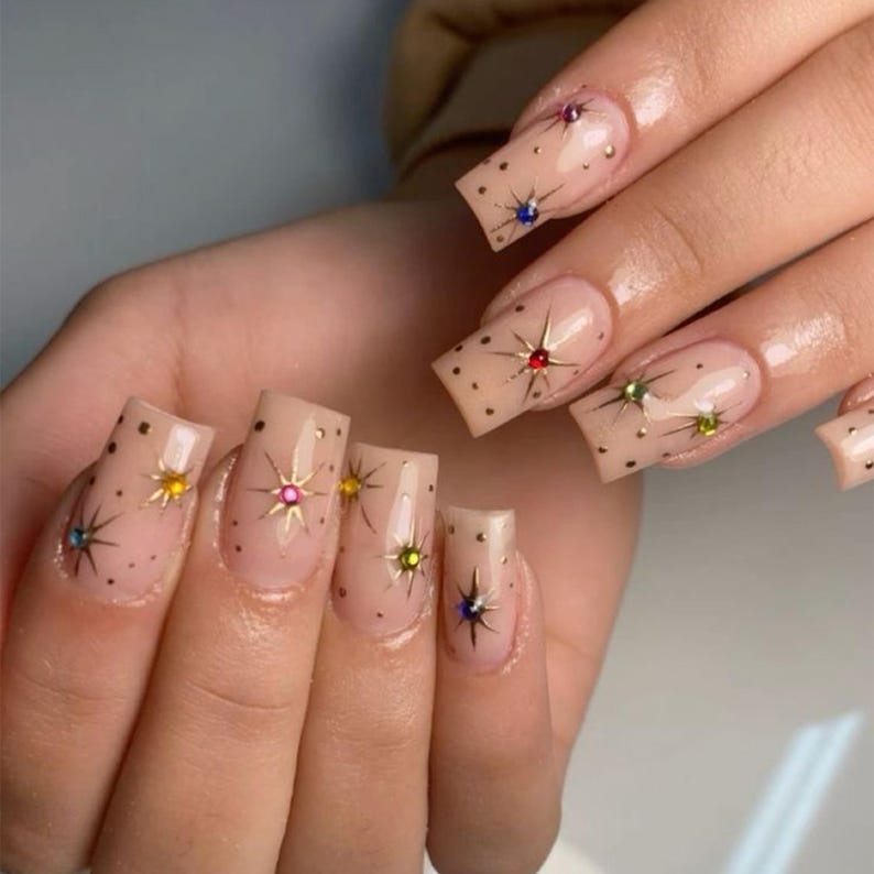 May include: A hand with square-shaped, light pink nails. Each nail features a gold starburst design with a colorful gem at the center. Small black dots are scattered across the nails, adding a playful touch to the manicure.