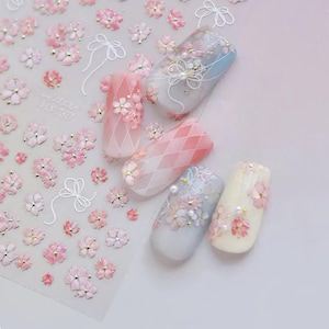 Könnte beinhalten: Ein Bogen mit Nagelaufklebern mit rosa Kirschblütenmotiven und weißen Linien-Schleifen, zusammen mit vier künstlichen Nagelspitzen, die mit passenden Blumenmustern, Rautenmustern und kleinen weißen Perlen verziert sind.