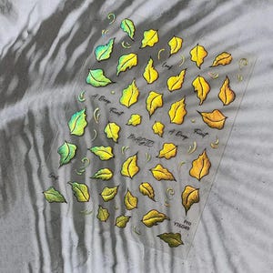May include: Clear sheet of nail art stickers featuring holographic leaf designs in shades of yellow, green, and gold. The words "Leaf", "A Deep Forest", and "Magic" are written on the sheet. The product code "YT6049" is also visible.