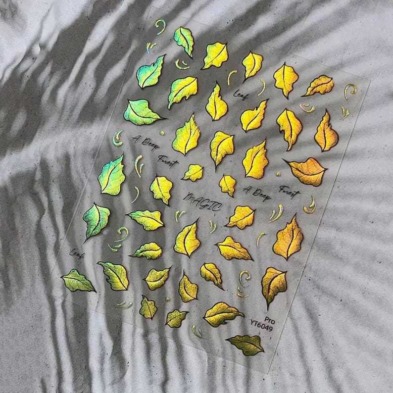 May include: Clear sheet of nail art stickers featuring holographic leaf designs in shades of yellow, green, and gold. The words "Leaf", "A Deep Forest", and "Magic" are written on the sheet. The product code "YT6049" is also visible.