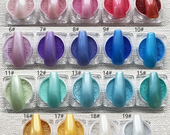 May include: A collection of 19 small, clear jars filled with shimmering nail powders in various colors. Each jar has a matching, long, oval-shaped nail painted on top. Colors range from pinks and purples to blues, greens, golds, and silvers.