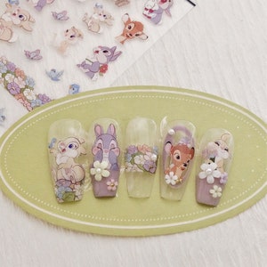 Peut inclure: Cinq pointes d'ongles transparentes avec des motifs floraux et des autocollants de personnages Disney. Les personnages incluent Bambi, Panpan et Miss Bunny. Les pointes sont sur un pr&eacute;sentoir ovale vert clair.