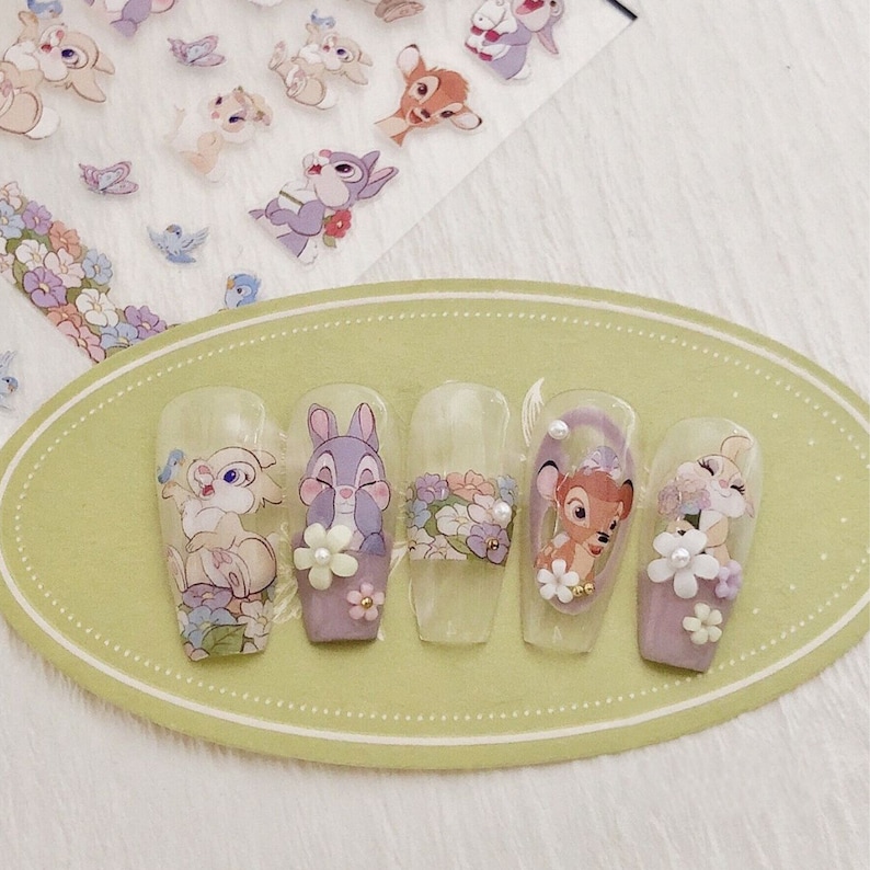 Peut inclure: Cinq pointes d'ongles transparentes avec des motifs floraux et des autocollants de personnages Disney. Les personnages incluent Bambi, Panpan et Miss Bunny. Les pointes sont sur un pr&eacute;sentoir ovale vert clair.