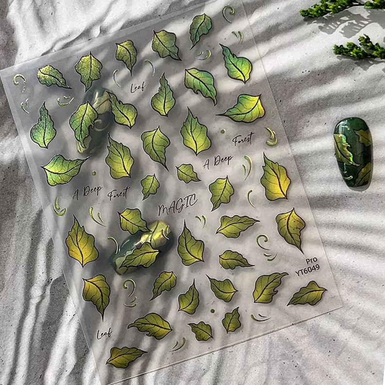 May include: Clear sheet of nail art stickers featuring green and yellow leaf designs. The words "Leaf", "A Deep Forest", and "Magic" are printed on the sheet. A painted nail with a leaf design is also visible.