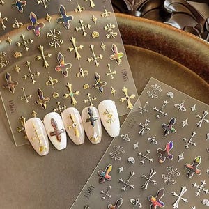 May include: Nail art stickers featuring gold and iridescent crosses, fleur-de-lis, and other decorative elements. Several nails are painted with white and nude polish, adorned with the stickers. The word "CROSS" is printed on the sticker sheets.