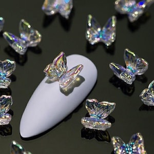 May include: Iridescent butterfly nail art decorations. These small, clear butterfly charms have a shimmering, multi-colored effect. One butterfly is placed on a white nail, surrounded by other butterfly charms. The background is a dark, reflective surface.
