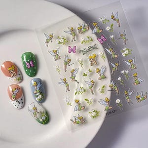 Op de afbeelding: Nagel stickers met Tinkerbell en vlinders. De stickers zijn op een witte achtergrond met groene bladeren en bloemen. De stickers zijn ontworpen om op nagels te worden geplakt.
