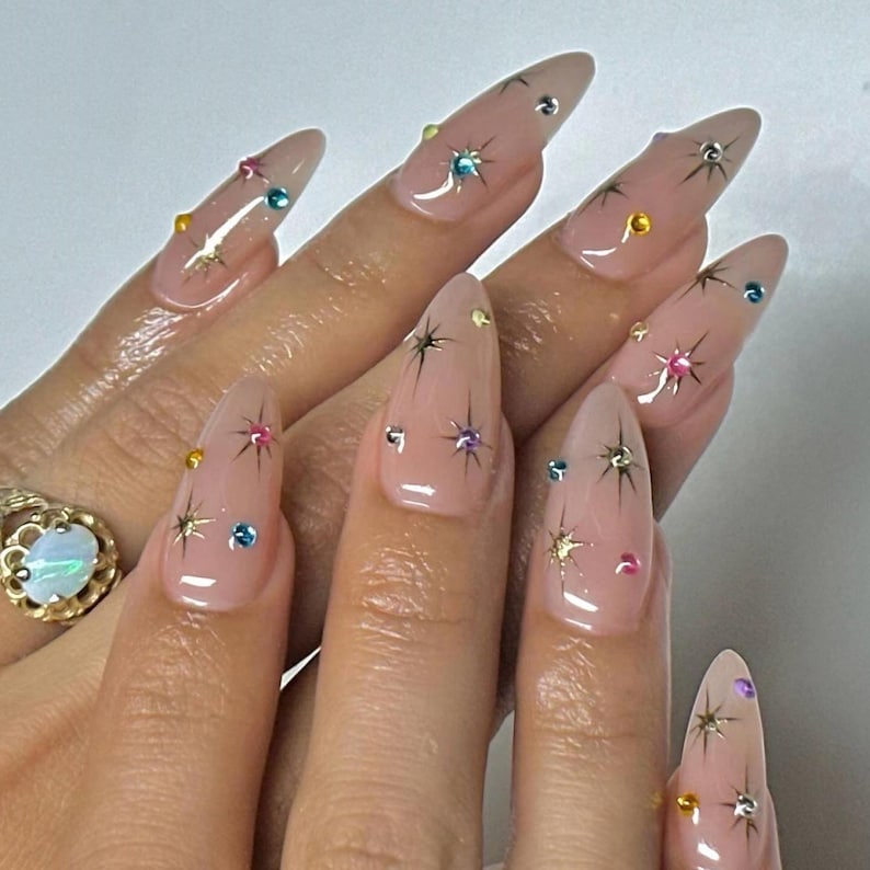 May include: Almond-shaped, light pink acrylic nails adorned with colorful rhinestones and starburst designs. The nails feature a clear top coat, enhancing the sparkle of the gems and the intricate star patterns.