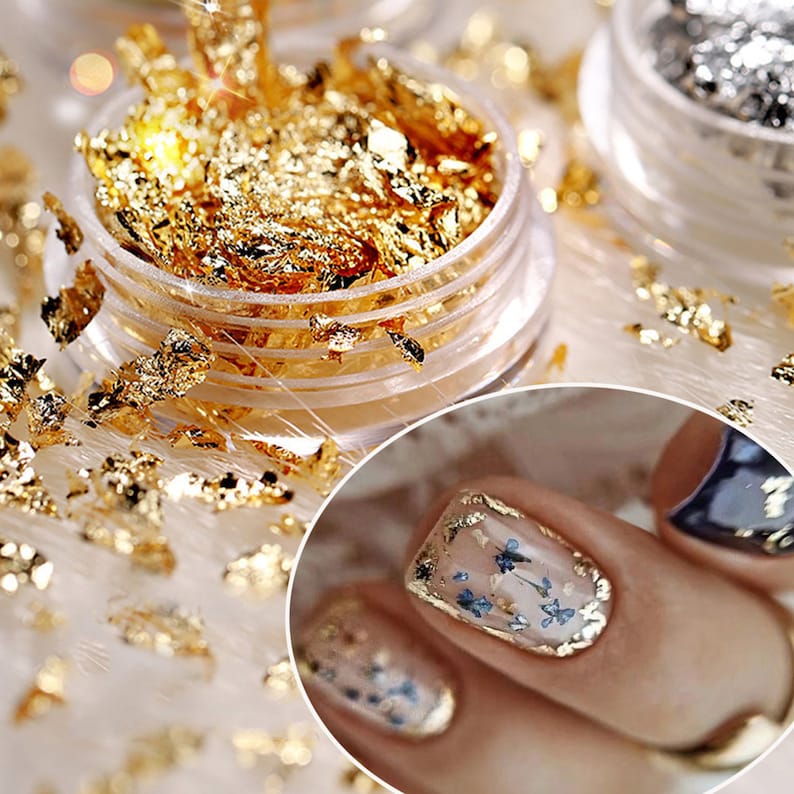 May include: A clear jar filled with gold leaf flakes. The jar is open and the flakes are spilling out. A close-up of a fingernail with a clear coat of nail polish and gold leaf flakes on top. The nail is decorated with small, dried flowers.