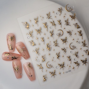 Peut inclure: Décalcomanies pour ongles en forme de fée dorée et de croissant de lune sur une feuille transparente. La feuille a un fond blanc et les décalcomanies sont dorées. La feuille comporte également de petites étoiles dorées et des strass. La feuille est étiquetée "Co-192".