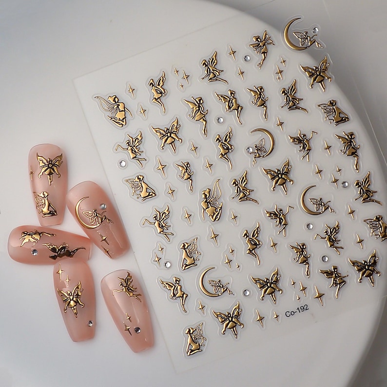May include: Gold fairy and crescent moon nail decals on a clear sheet. The sheet has a white background and the decals are gold. The sheet also has small gold stars and rhinestones. The sheet is labeled "Co-192".