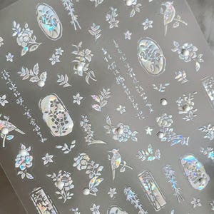 Puede incluir: L&aacute;mina transparente de pegatinas iridiscentes para decoraci&oacute;n de u&ntilde;as con dise&ntilde;os florales, p&aacute;jaros y texto japon&eacute;s. Las pegatinas incluyen varias formas de flores, hojas y peque&ntilde;os acentos perlados. El dise&ntilde;o general es delicado y detallado, con un efecto hologr&aacute;fico.