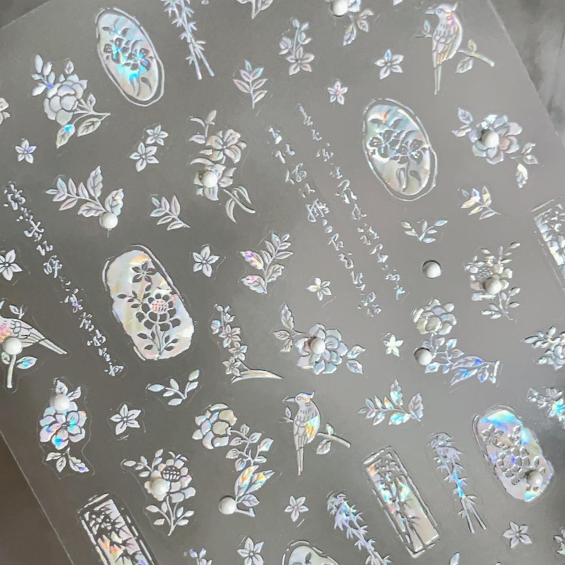 Puede incluir: L&aacute;mina transparente de pegatinas iridiscentes para decoraci&oacute;n de u&ntilde;as con dise&ntilde;os florales, p&aacute;jaros y texto japon&eacute;s. Las pegatinas incluyen varias formas de flores, hojas y peque&ntilde;os acentos perlados. El dise&ntilde;o general es delicado y detallado, con un efecto hologr&aacute;fico.