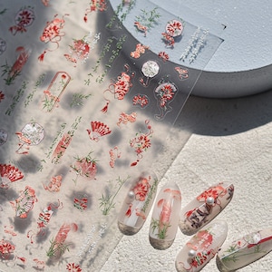 K&ouml;nnte beinhalten: Eine Blatt mit transparenten Nagelaufklebern mit einem floralen und Bambus-Design in Rot, Gr&uuml;n und Wei&szlig;. Die Aufkleber sind auf f&uuml;nf nackten Fingern&auml;geln abgebildet.