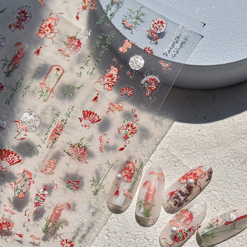 K&ouml;nnte beinhalten: Eine Blatt mit transparenten Nagelaufklebern mit einem floralen und Bambus-Design in Rot, Gr&uuml;n und Wei&szlig;. Die Aufkleber sind auf f&uuml;nf nackten Fingern&auml;geln abgebildet.