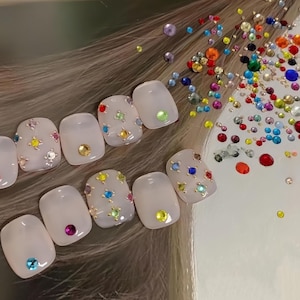 May include: Close-up of ten white, square-shaped fake nails with colorful rhinestones arranged in a starburst pattern on each nail.