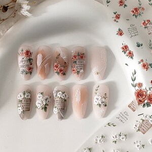 Puede incluir: U&ntilde;as artificiales con dise&ntilde;os florales. Las u&ntilde;as presentan rosas rojas y blancas, escritura y detalles de perlas. Algunas u&ntilde;as tienen una base nude con l&iacute;neas blancas. Tambi&eacute;n se ve una hoja de arte de u&ntilde;as floral similar.