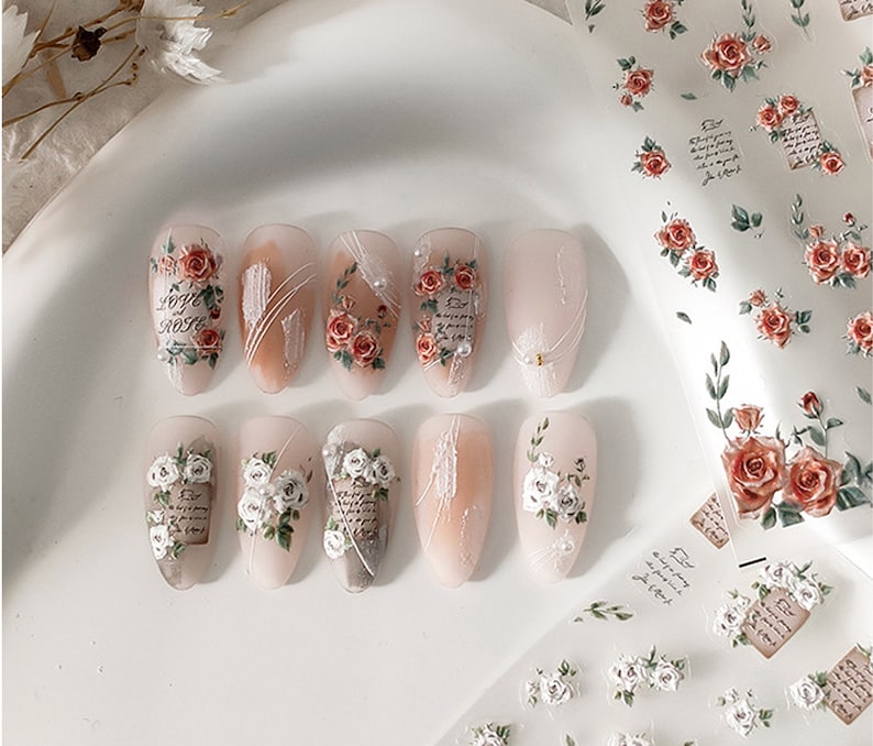 Puede incluir: U&ntilde;as artificiales con dise&ntilde;os florales. Las u&ntilde;as presentan rosas rojas y blancas, escritura y detalles de perlas. Algunas u&ntilde;as tienen una base nude con l&iacute;neas blancas. Tambi&eacute;n se ve una hoja de arte de u&ntilde;as floral similar.