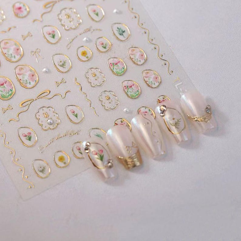 May include: A sheet of nail art stickers featuring floral and decorative designs in gold, pink, and white. The stickers include flowers, bows, and pearl accents. Several manicured nails are shown with similar designs.