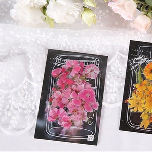 May include: A set of two clear glass jar stickers with pressed flowers. One sticker features pink flowers and the other features yellow flowers. Both stickers have a black background and white outlines.