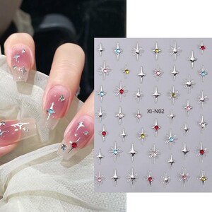 May include: A sheet of nail art stickers featuring silver starburst designs with colorful gem centers. The image also shows a hand with long, clear acrylic nails decorated with similar starburst gems in various colors.