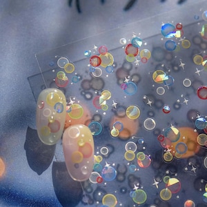 May include: A clear sheet of nail art stickers featuring a pattern of colourful translucent circles and small four-pointed stars. Two oval-shaped artificial fingernails are displayed on the left, each decorated with matching circular designs.