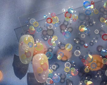 May include: A clear sheet of nail art stickers featuring a pattern of colorful translucent circles and small four-pointed stars. Two oval-shaped artificial fingernails are displayed on the left, each decorated with matching circular designs.