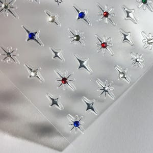 May include: Sheet of silver star-shaped nail art stickers. Each star has a central gem in various colors, including red, blue, and clear. The stickers are on a clear, transparent backing.