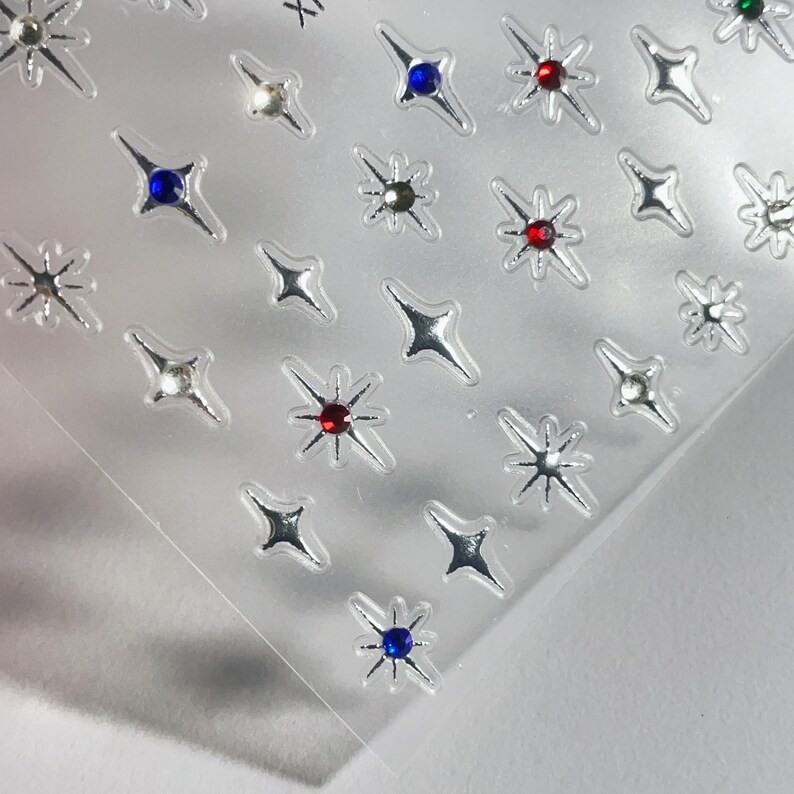 May include: Sheet of silver star-shaped nail art stickers. Each star has a central gem in various colors, including red, blue, and clear. The stickers are on a clear, transparent backing.
