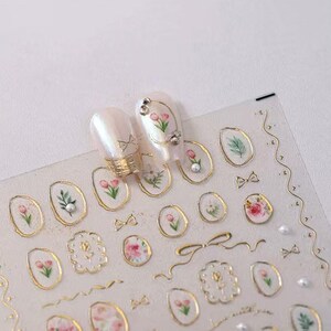 May include: Clear nail art stickers with gold trim and floral designs. The stickers feature small pink tulips, green leaves, bows, and pearls. Two nails are decorated with the stickers, showcasing the design.
