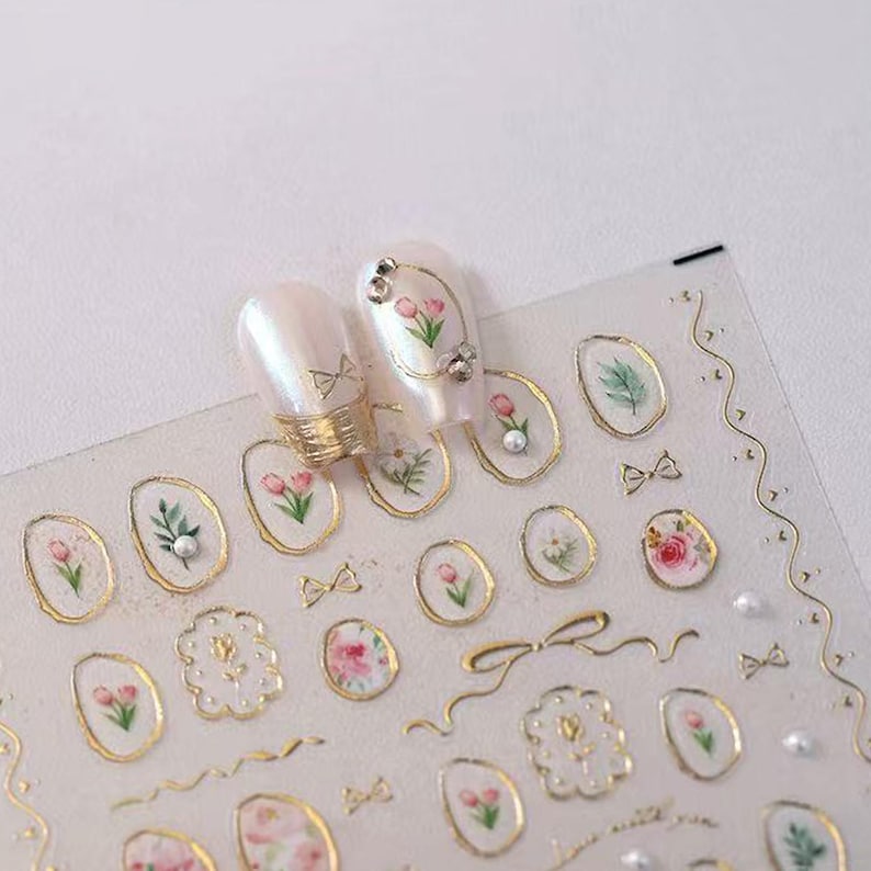 May include: Clear nail art stickers with gold trim and floral designs. The stickers feature small pink tulips, green leaves, bows, and pearls. Two nails are decorated with the stickers, showcasing the design.
