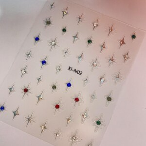 May include: Clear sheet of nail art stickers with various star and gemstone designs. The stickers feature silver, red, green, and blue gems, and the text "XI-N02". These stickers are perfect for adding a touch of sparkle to any manicure.