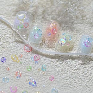 May include: Close-up of a set of manicured nails with a bubble-themed design. The nails are painted in a clear, glittery polish with colorful, iridescent bubble accents. Additional bubble-shaped embellishments are scattered around the nails.