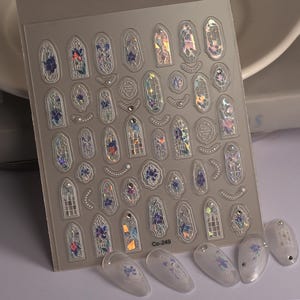 May include: A sheet of clear nail stickers featuring a stained glass window design. The stickers are iridescent and come in a range of colours, including blue, purple, and pink. The sheet also includes the text "Co-249".