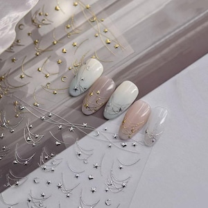 Può includere: Adesivi per nail art con disegni celesti dorati e argentati. Gli adesivi includono stelle, lune crescenti e linee curve. Diverse unghie curate sono decorate con gli adesivi.