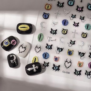 May include: Nail art stickers featuring various designs, including cat eyes, crosses, crescent moons, and flame hearts. The stickers come in black, white, and silver, with accents of glitter and the text "The Devil" and "Co-334".