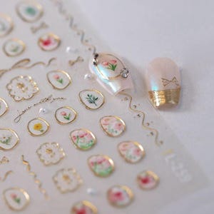 May include: A sheet of nail art stickers featuring floral designs and gold accents. The stickers include various flower illustrations, gold borders, and the text "Love with you." Two manicured nails are also shown, one with a floral design and the other with a gold base.